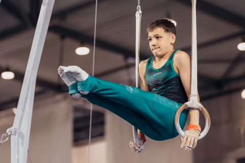 Young male gymnast holds still rings with concentration, legs raised mid-routine in an indoor gymnastics hall.