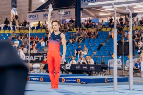 Male gymnast raises hand to acknowledge crowd after parallel bars routine at Sport Vlaanderen competition.