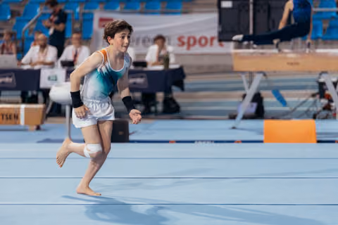 Young male gymnast sprints barefoot across the blue floor, focused and determined, during a gymnastics competition.