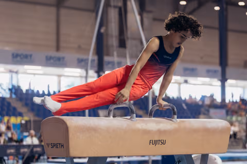 Young male gymnast with curly hair concentrates intensely while performing on the pommel horse at an indoor competition.