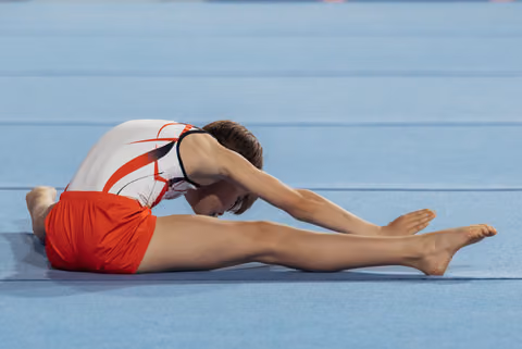 Young male gymnast in red and white leotard performs a seated forward stretch on a blue competition floor, head bowed.
