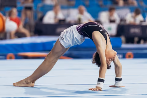 Young gymnast performs a deep backbend on the floor exercise mat during a competition, barefoot with wrist guards.