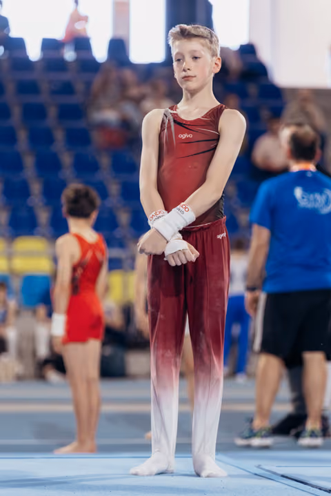Young male gymnast in dark red agiva leotard stands composed, wrists wrapped, awaiting his turn on the competition floor.