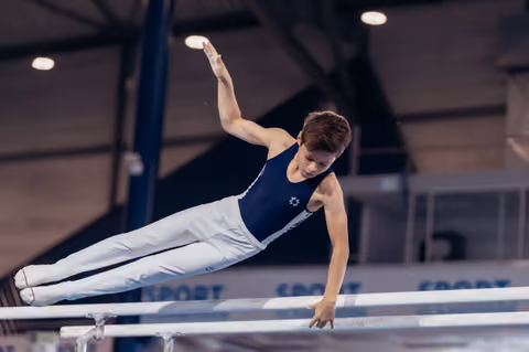 Young male gymnast performs a precise horizontal hold on parallel bars, one arm raised, showing intense focus.