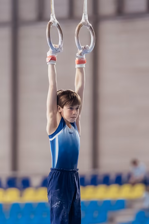 Focused young male gymnast hangs from still rings, arms raised overhead, wearing blue leo in a sports hall.