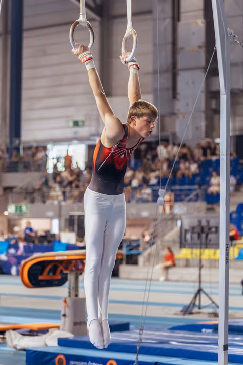 Young male gymnast hangs from still rings with focused expression, arms fully extended, in a busy sports hall.