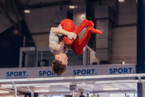 Young male gymnast upside-down in a tucked somersault above parallel bars, smiling mid-flight in an indoor sports hall.