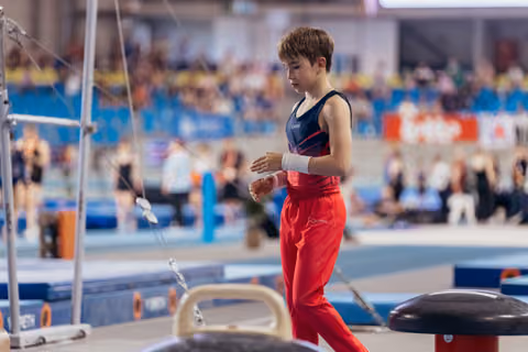 Young male gymnast in red and navy leotard applies chalk to his hands, focused and composed before his routine.