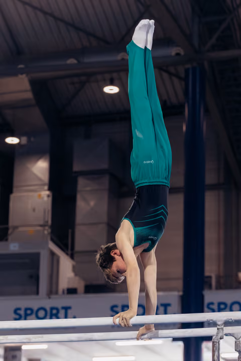 Young male gymnast holds a perfect handstand on parallel bars, legs straight up, wearing teal outfit in indoor gym.