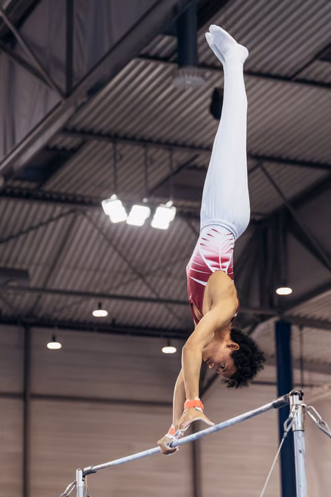 Male gymnast performs a perfect handstand on the horizontal bar, body fully extended, legs pointing skyward in an indoor arena.