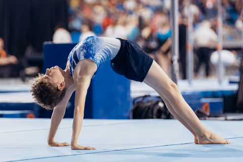 Young male gymnast performs a back bridge on the floor exercise mat, showcasing flexibility and focus during a gymnastics meet.