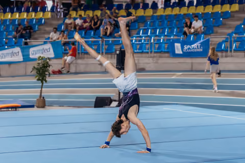 Young male gymnast performs a one-armed handstand on the blue floor, legs split wide, at a Gymfed indoor competition.