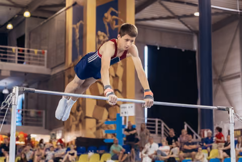 Determined teenage male gymnast grips the high bar mid-routine, body horizontal, intense focus on his face, indoor arena crowd behind.