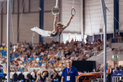 Young male gymnast holds a cross position on still rings, intense focus, crowd-filled arena in background.