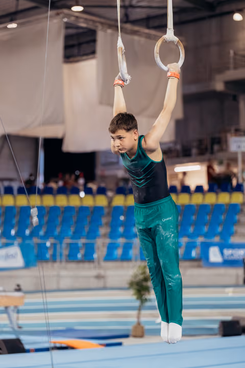 Young male gymnast hangs from still rings with intense focus, wearing green and black leotard in a competition arena.