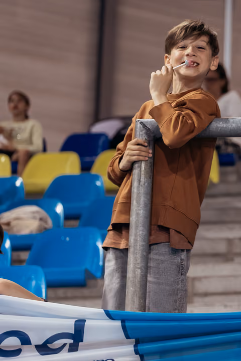 Young boy grins while eating a lollipop, leaning on a metal railing in colorful stadium bleachers.