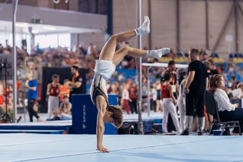 Young male gymnast performing a one-arm handstand on the floor exercise mat during a packed competition event.