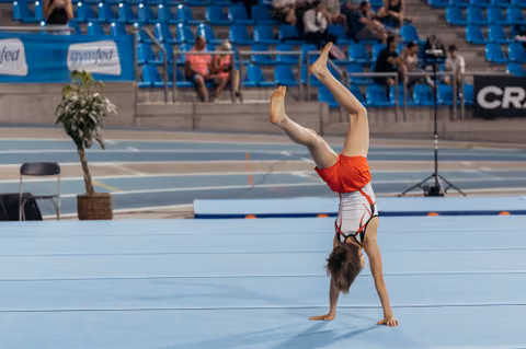Young gymnast performs a one-handed cartwheel on the blue competition floor, legs split wide in a gymnastics arena.