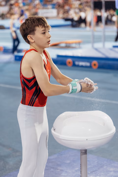 Young male gymnast applies chalk to his hands with focused concentration, wearing red and black leotard at a competition arena.