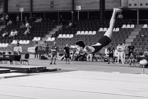 Male gymnast mid-backflip over the floor mat, body fully extended, during an indoor gymnastics competition.
