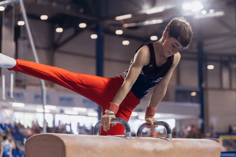 Young male gymnast performs on pommel horse, legs extended sideways, focused expression, in a large indoor gymnastics hall.