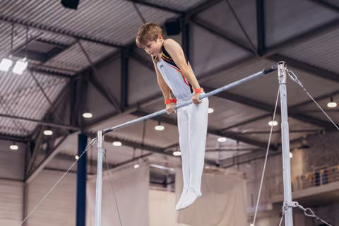 Young male gymnast grips the high bar with intense focus, body extended, competing in a large indoor gymnasium.