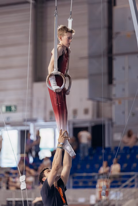 Young male gymnast hangs from still rings with intense focus while a coach supports his feet below in a sports hall.