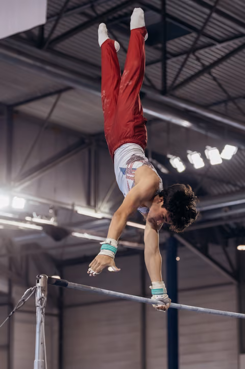 Young male gymnast performs an inverted giant on the high bar, body fully extended, in an indoor gymnastics hall.