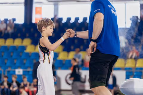 Young gymnast in white uniform fist-bumps coach in blue shirt, sharing a moment of encouragement in a busy sports arena.