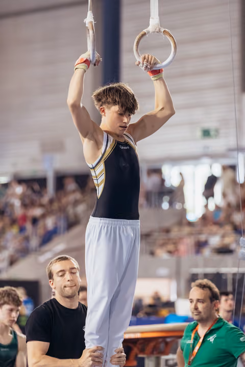 Young male gymnast hangs from still rings with intense focus, supported by coach below in a packed competition arena.