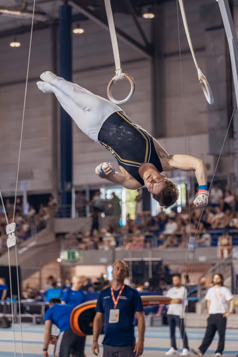 Male gymnast hangs inverted on still rings, face tense with effort, while a coach watches below in a packed arena.