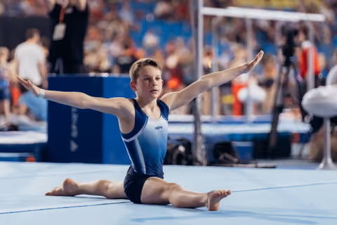 Young male gymnast performs a split on the floor with arms outstretched, displaying calm focus during a gymnastics event.