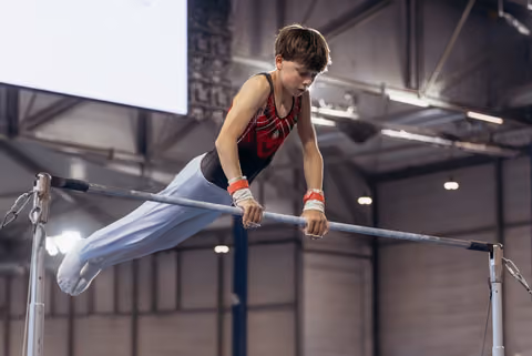 Determined young male gymnast grips the horizontal bar with intense focus, body extended mid-routine in an indoor gymnasium.