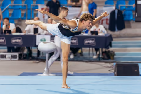 Young male gymnast holds a precise arabesque on the floor, arms extended, focused expression during a gymnastics meet.