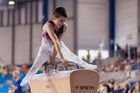 Young male gymnast performs on pommel horse, leaning forward with intense focus during a gymnastics meet.