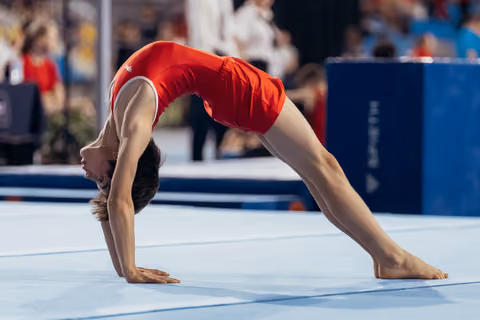 Young gymnast in red leotard performs a bridge pose on the floor exercise mat during a gymnastics competition.
