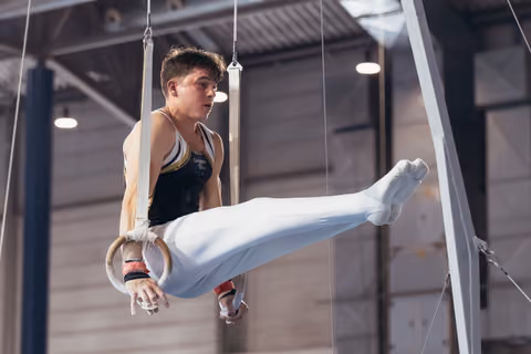 Young male gymnast holds still rings with intense focus, executing a precise leg raise in an indoor gymnasium.