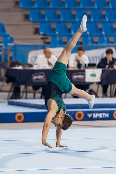 Young male gymnast performs a one-armed handstand with legs split on the floor exercise mat during competition.