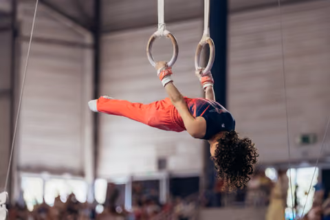 Young gymnast performs a dramatic iron cross hold on still rings, body fully inverted, showing strength and focus.