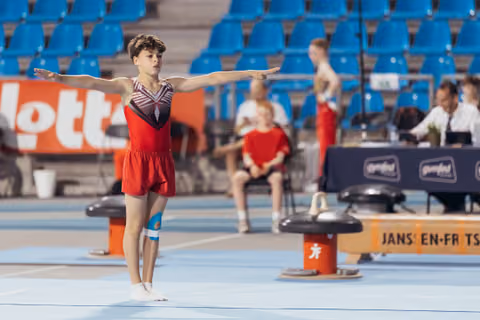 Young male gymnast performing floor exercise, arms outstretched with focused expression, in a competition arena with blue seats.