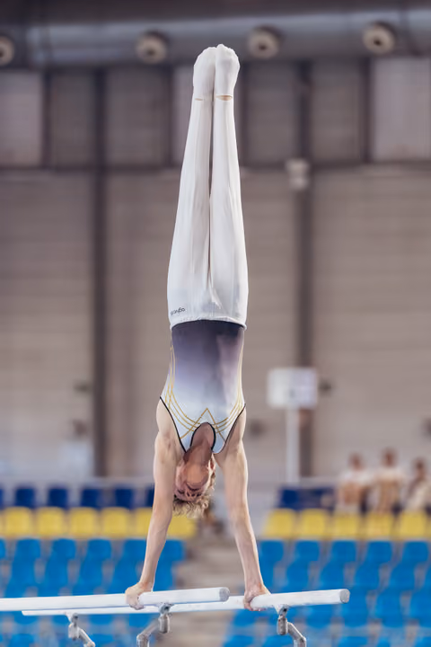 Young male gymnast holds a perfect handstand on parallel bars, body fully extended, in a competition hall.