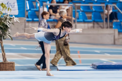 Young gymnast holds a precise arabesque balance on floor, arms extended, focused expression during competition.