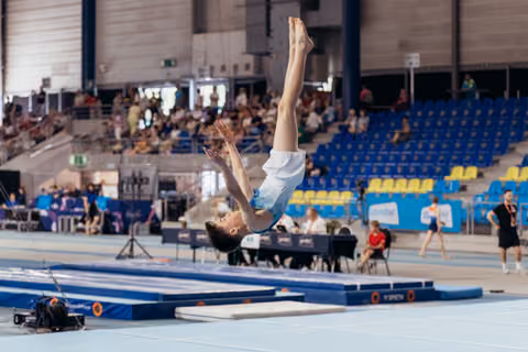 Young gymnast airborne in a back layout, legs extended skyward, competing in a blue-floored arena with spectators watching.