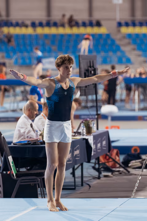 Young male gymnast stands with arms wide open on the floor mat, focused and composed, at an indoor gymnastics event.