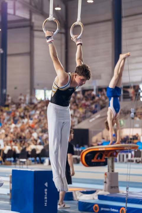 Young male gymnast grips still rings with intense focus, white trousers, packed arena crowd in background.