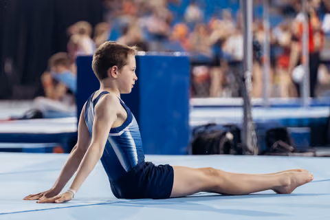 Focused young male gymnast performs a seated stretch on the floor exercise mat during a gymnastics meet.