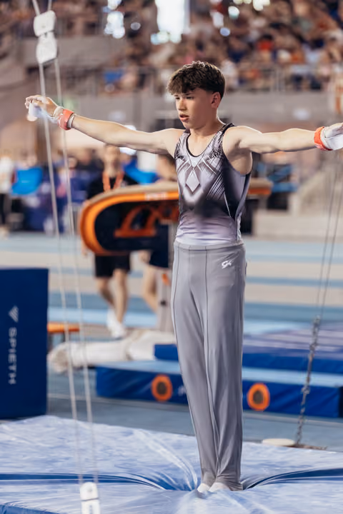 Young male gymnast stands with arms wide, focused expression, balancing on mat at indoor gymnastics competition.