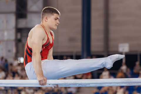 Young male gymnast performs an L-sit on parallel bars, focused expression, wearing red leotard and white pants.