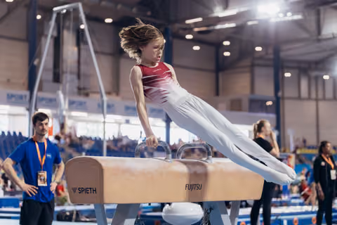 Young gymnast performs on pommel horse with fierce concentration, body parallel, in a busy sports hall.