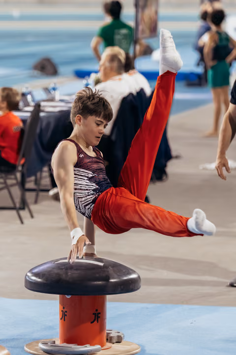 Young male gymnast performs a scissor element on the pommel horse, legs split high, expression calm and concentrated.